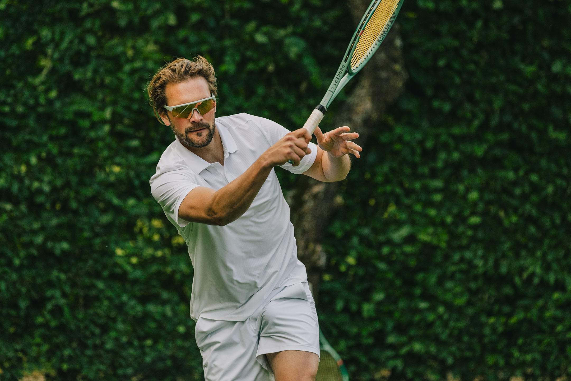 The ACE Hub Johnny Loco sport glasses with mirrored lenses, worn during a tennis match on a grass court.
