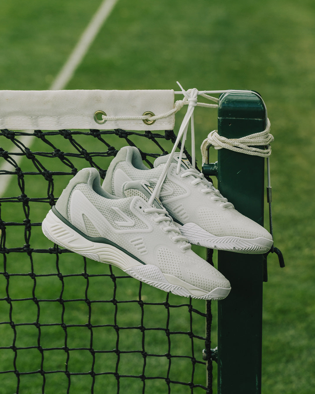 The ACE Hub Johnny Loco performance tennis shoes in off-white and cucumber, hanging on a grass court net.