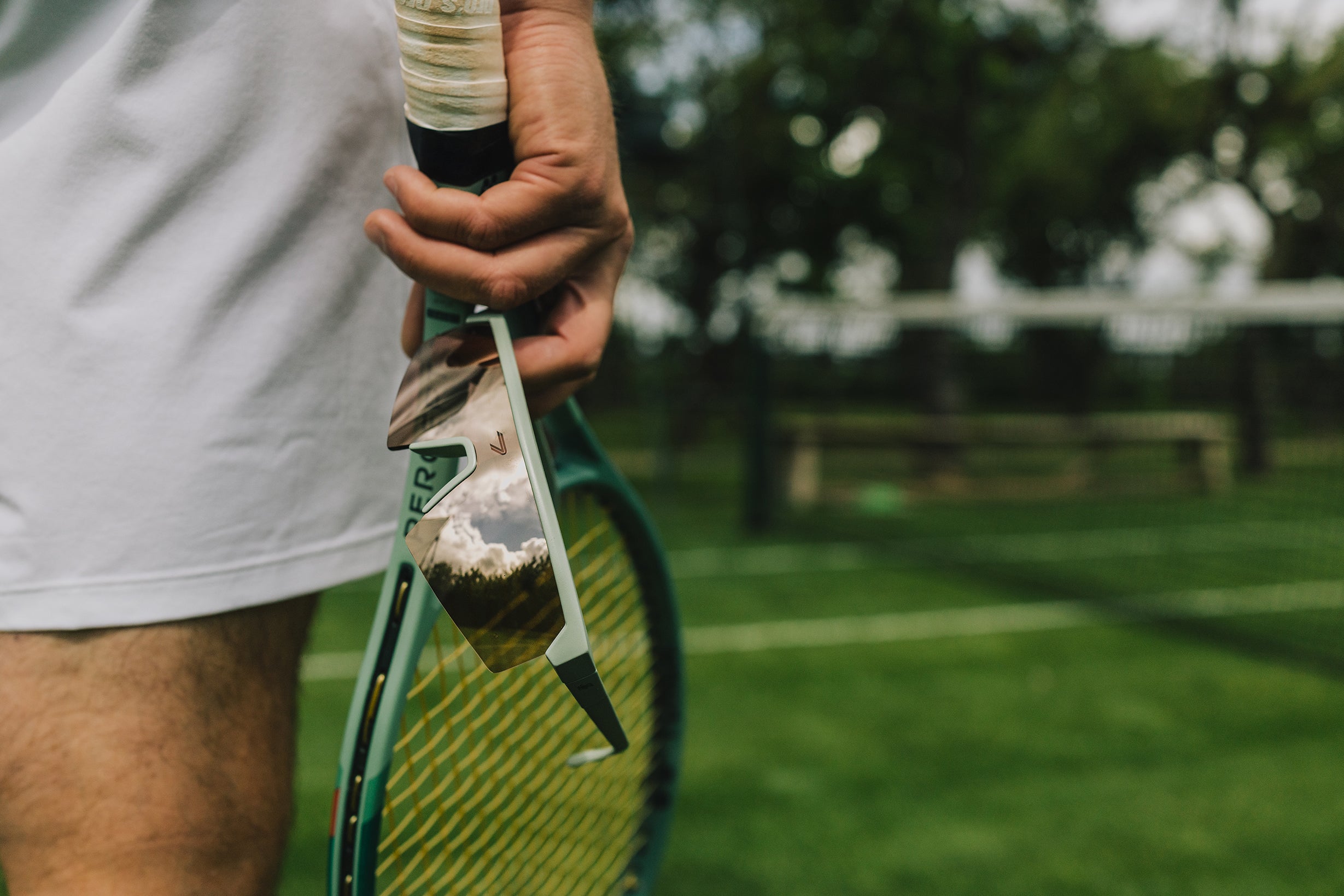 The ACE Hub Johnny Loco sport glasses with mirrored lenses, held on a grass tennis court.