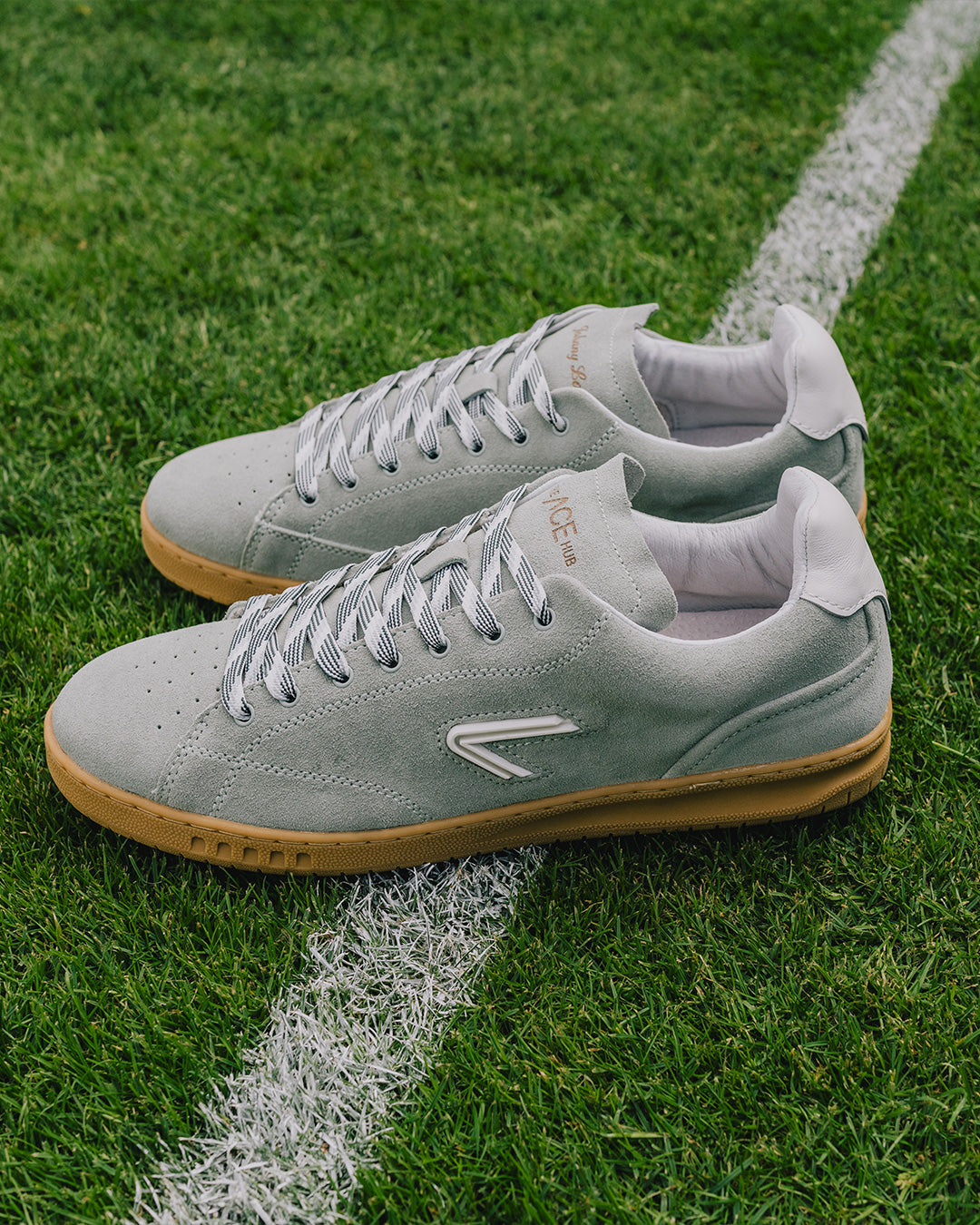 The ACE Hub Shot Suede sneaker in sage green, Johnny Loco collab edition, photographed on a grass tennis court.