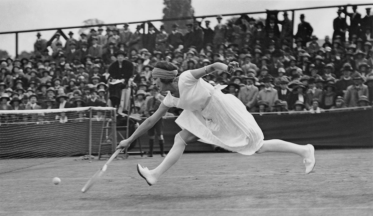 Black and white historical photograph of Suzanne Lenglen playing tennis on an outdoor court, captured mid-swing with a racket while wearing a white dress and headband, with a large crowd seated in the stands behind her.