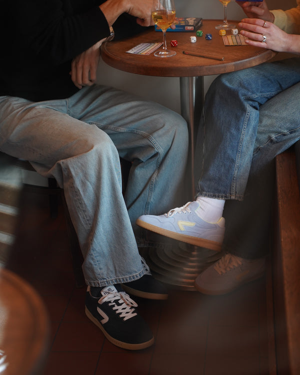 Two people sitting at a table with drinks and cards, wearing jeans and suede hub sneaker.