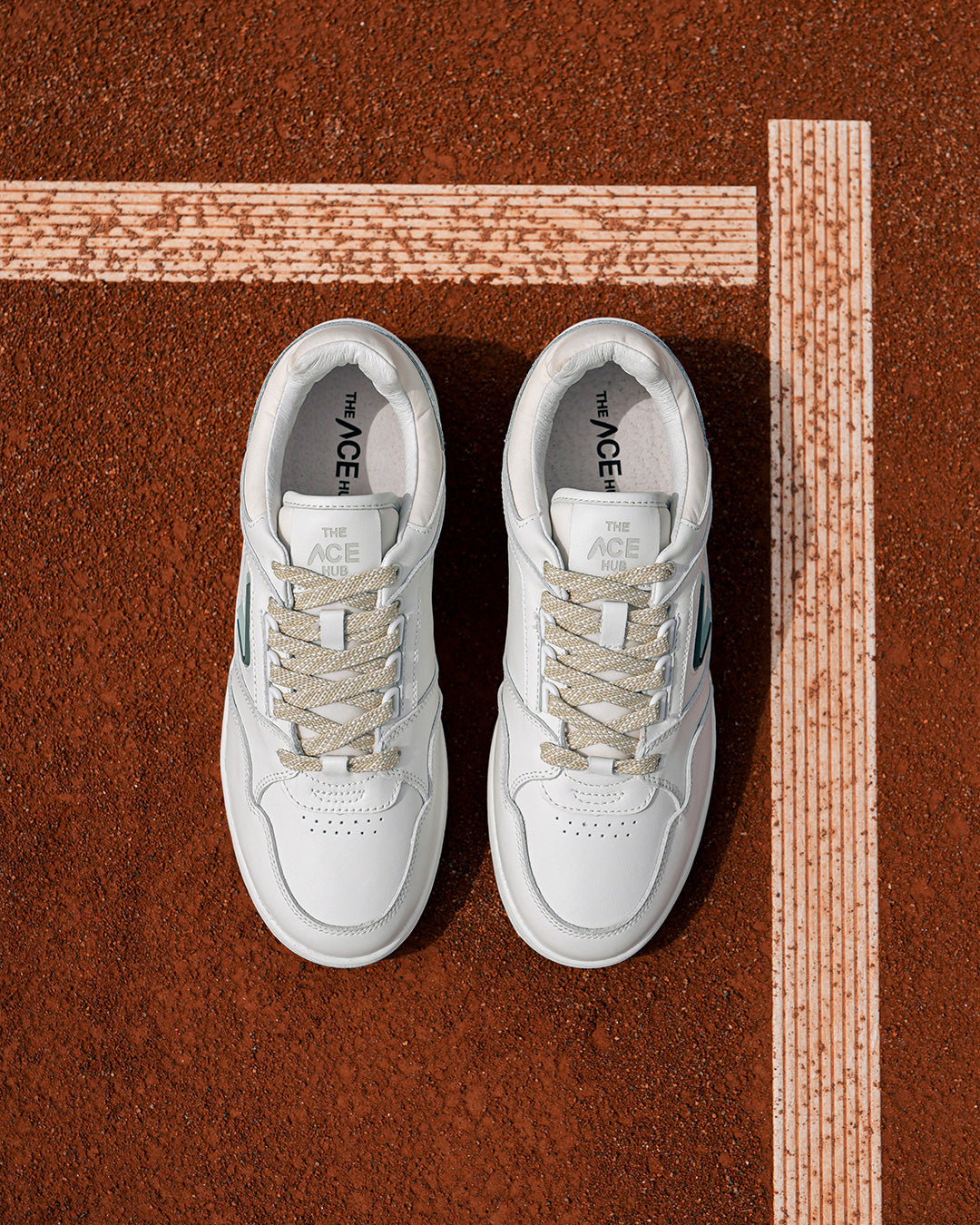 The ACE Hub Sprint sneaker in off-white and cucumber, photographed on a clay tennis court.