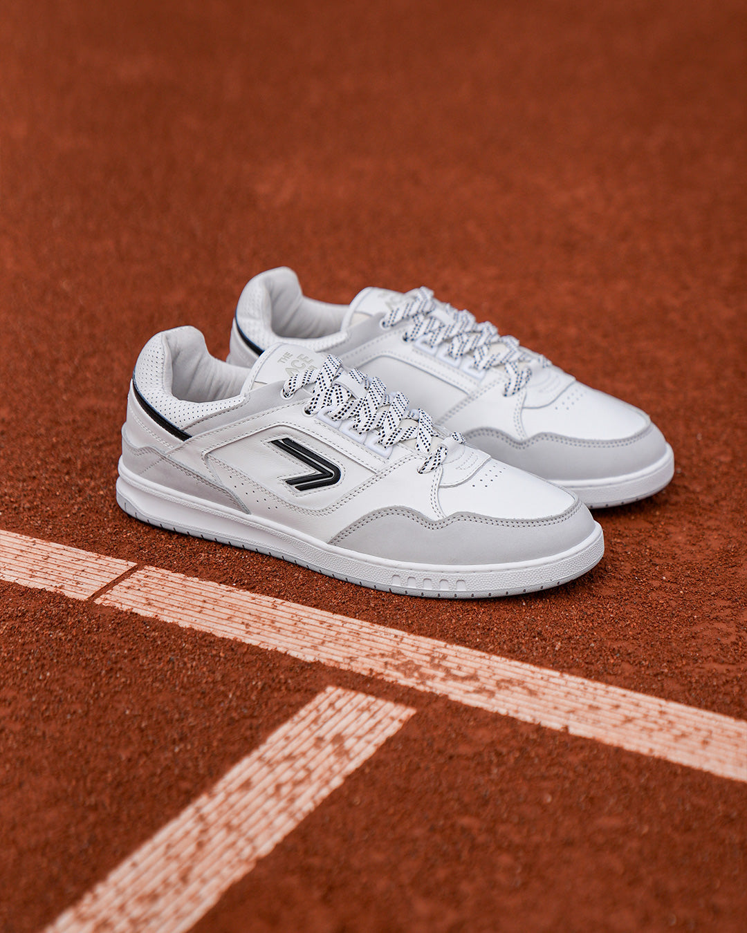 The ACE Hub Sprint sneaker in white and black, photographed on a clay tennis court.
