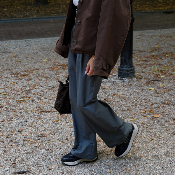 Person wearing a brown coat, gray pants and black sneakers walking on a gravel path.